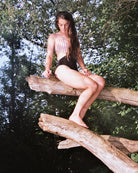 Im Badeanzug Luna von Main Design mit seitlichen Ausschnitten sitzt eine Frau auf einem großen Ast über dem Wasser inmitten von üppigem Grün und natürlichem Licht und blickt entspannt nach unten.