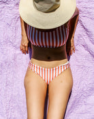 Eine Frau in einer Luxe Bikini Hose von Main Design, mit brasilianischem Schnitt und rot-weißen Streifen, entspannt sich auf einem lavendelfarbenen Handtuch. Ihr Gesicht wird von einem breitkrempigen Strohhut verdeckt. Einige Stücke sind aus recycelten Materialien hergestellt. Das Foto ist von oben aufgenommen.