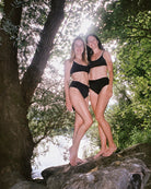 Zwei Frauen in schwarzen Badeanzügen stehen barfuß auf Felsen an einem Fluss, umgeben von Bäumen und Sonnenlicht, das durch grünes Laub fällt.