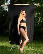 Eine Frau trägt das Sassy Bikini-Top von Main Design und steht barfuß im Gras im Sonnenlicht vor einem schwarzen Stoffhintergrund im Freien, umgeben von grünen Bäumen und Blättern.