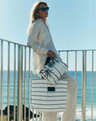 Eine Frau mit Sonnenbrille und heller Kleidung steht an einem Metallgeländer und hält eine gestreifte Tragetasche sowie die „Aloha Collection Aloha Mid Pouch“ mit Palmenmuster in der Hand. Im Hintergrund sind das Meer und der blaue Himmel zu sehen.