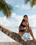 Eine Frau in einem schwarzen Bikini sitzt auf einer Palme über einem tropischen Strand und hält die „Aloha Collection Aloha Mid Pouch“ – einen schicken Reiseorganizer mit blau-weißem Blumenmuster – in der Hand, während eine weiße Blume in ihrem Haar steckt.