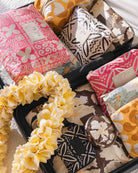 Ein Koffer voller bunter, gemusterter Beutel – darunter der „Aloha Small Pouch“ von Aloha Collection, eine wasserabweisende Kosmetiktasche – sowie ein gelber Blumen-Lei und eine runde, gemusterte Puderdose, alles ordentlich auf einem Stoff mit tropischem Muster angeordnet.
