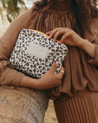Eine Person in brauner Kleidung steht neben einer Palme und hält einen „Aloha Collection Aloha Small Pouch“ in der Hand – eine Reißverschlusstasche mit Leopardenmuster und einem weißen „ALOHA“-Etikett.