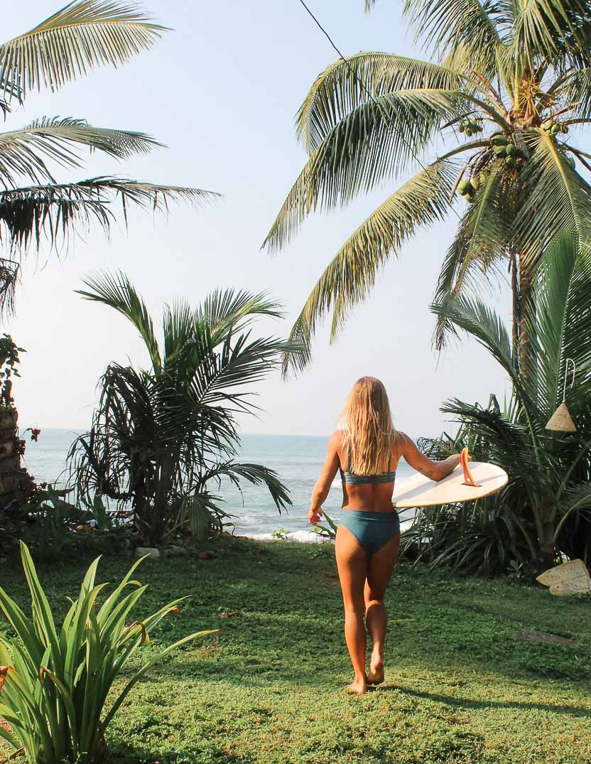 Eine Frau mit langen Haaren läuft barfuß über eine Rasenfläche in Richtung Meer und hält ein Surfbrett unter dem Arm. Sie trägt ein blaugrünes Sassy Bikini Oberteil von Main Design und die Szene wird von Palmen und anderen tropischen Pflanzen unter einem klaren Himmel eingerahmt.