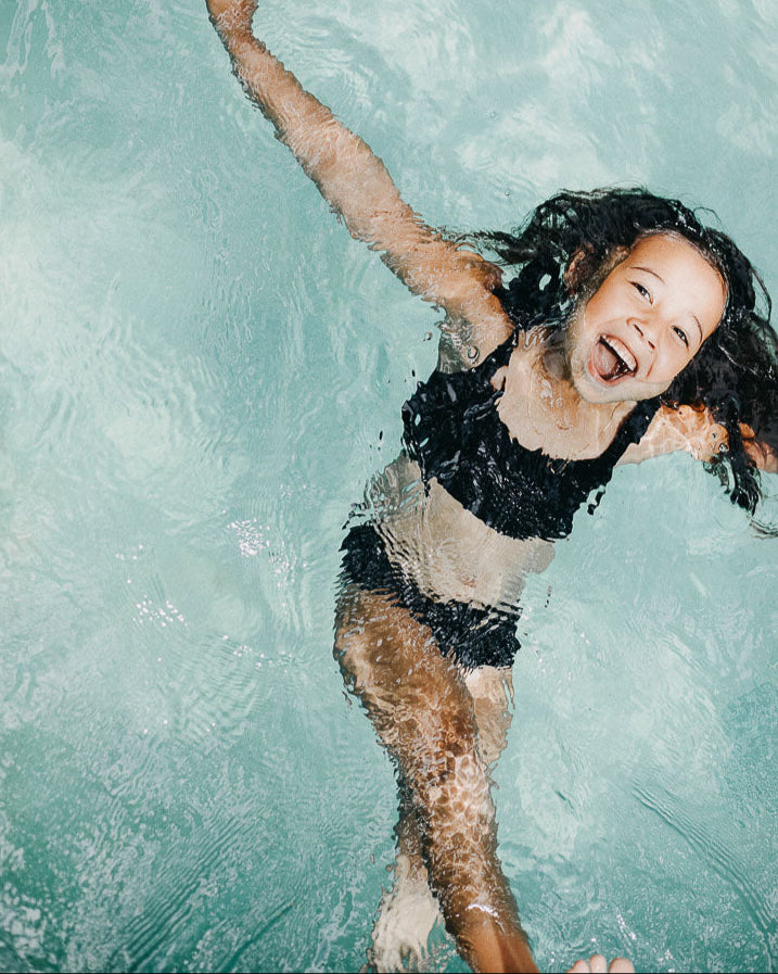 Ein junges Mädchen im schwarzen Mia Mädchen Bikini von Main Design schwimmt mit ausgestreckten Armen auf dem Rücken in einem Schwimmbecken und lächelt fröhlich. Ihr langes, dunkles Haar liegt ausgebreitet im Wasser und sie wirkt entspannt und glücklich. Das Wasser ist klar und blau.