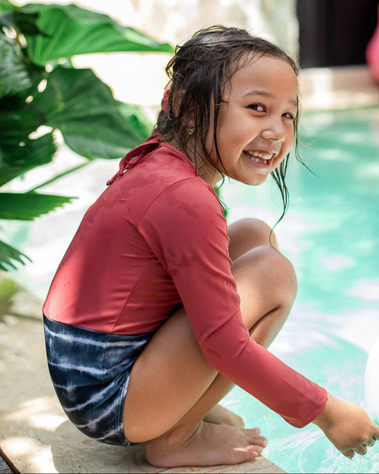 Ein kleines Kind mit nassen Haaren, das einen Laila Mädchen Badeanzug von Main Design und blaue Badeshorts trägt, hockt am Rand eines Pools und lächelt strahlend in die Kamera. Üppiges grünes Laub im Hintergrund verstärkt die fröhliche und lebendige Szene.