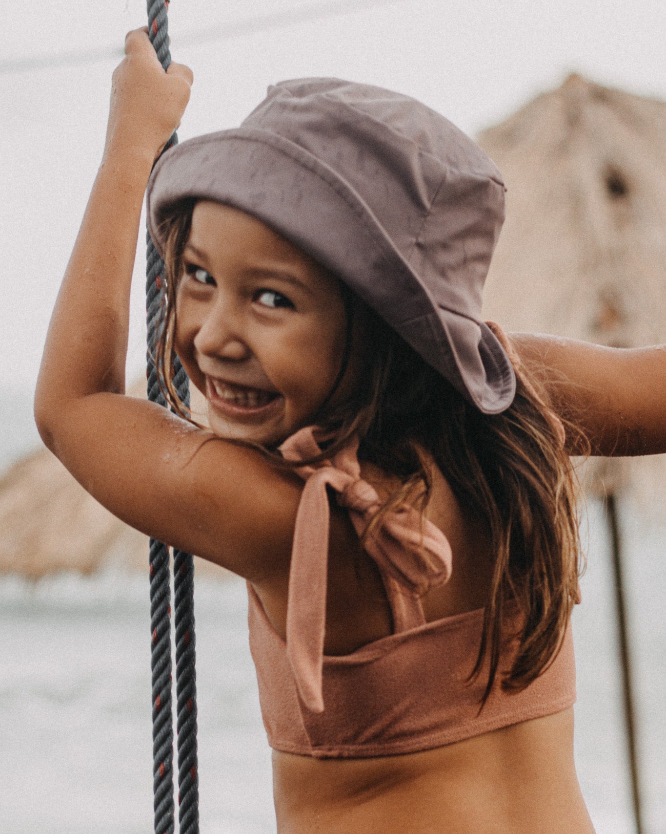 Ein junges Mädchen in einem hellbraunen Bikini und einem Kinder-Sonnenhut von Main Design lächelt und hält ein Seil. Sie steht an einem sonnigen Strand mit dem Meer und den Felsen im Hintergrund und genießt den Sonnenschutz UPF 50+.