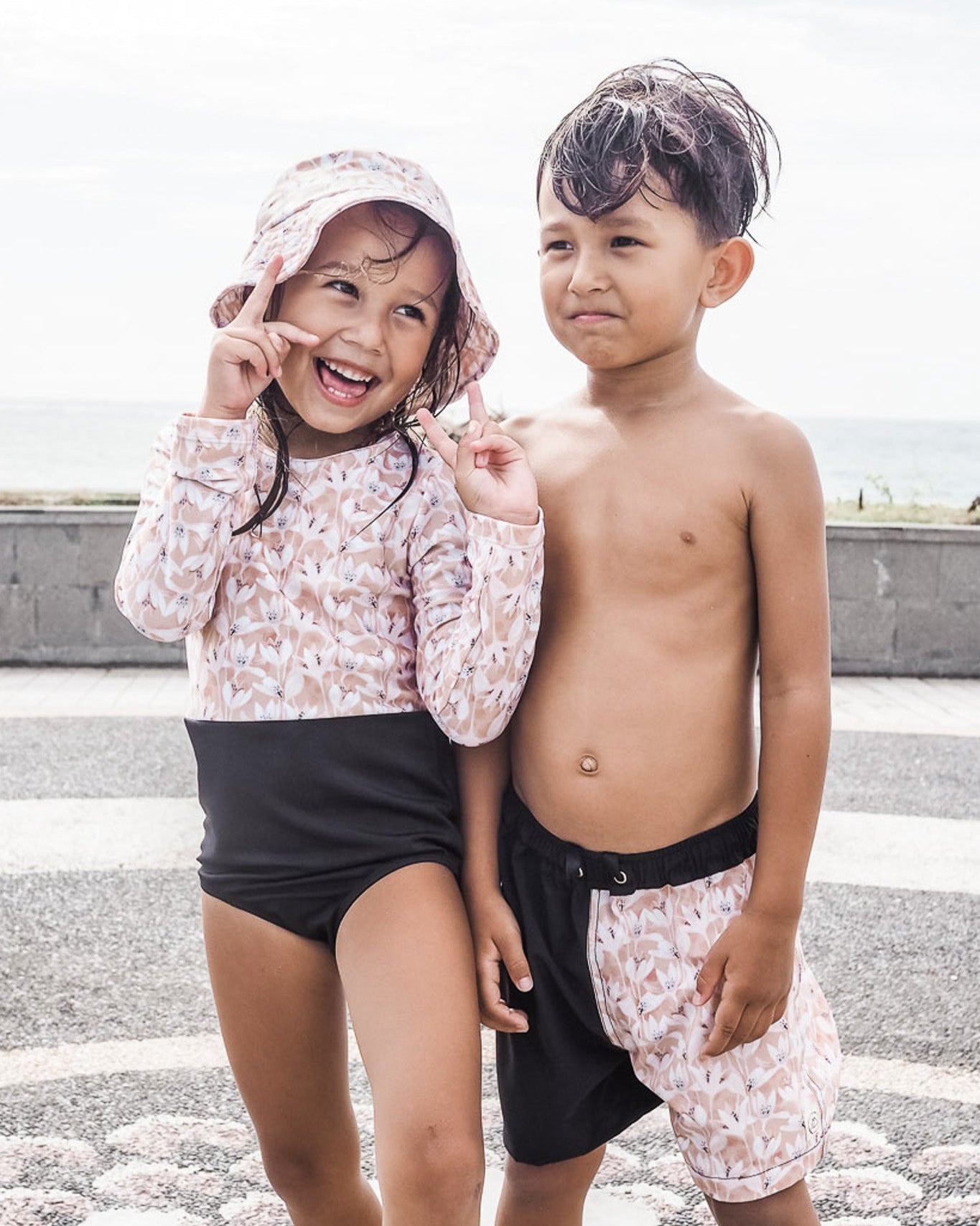 Ein junges Mädchen und ein junger Junge, beide in Bademode von Main Design, stehen am Strand. Das Mädchen strahlt in ihrem Rashguard mit LSF 50+ und Shorts, während der Junge passende Badeshorts trägt. Ihr gemeinsamer Moment wird durch den ruhigen Ozean im Hintergrund eingerahmt.