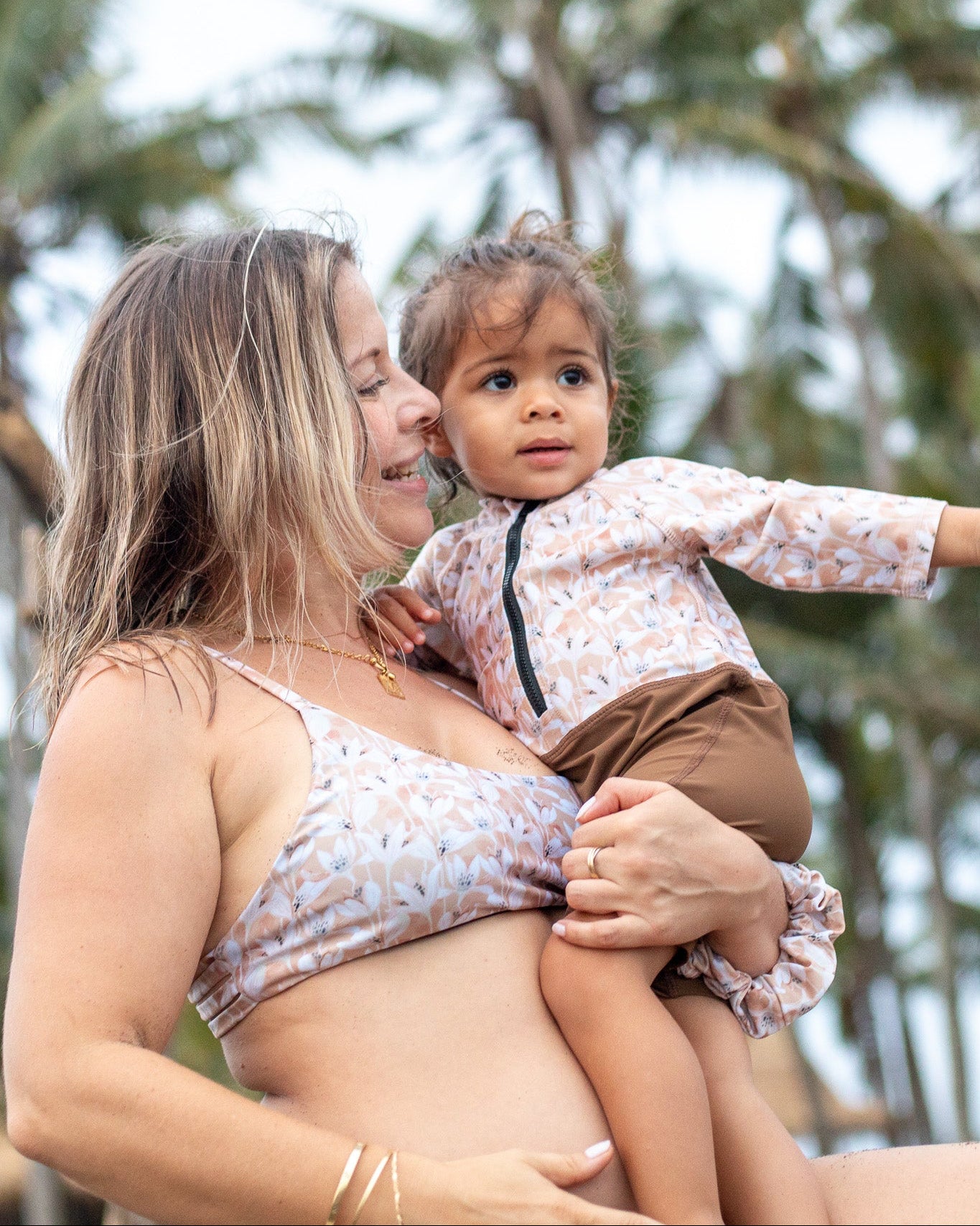 Eine Frau und ihr Kleinkind genießen einen nachhaltigen Strandtag unter Palmen und tragen passende schnelltrocknende Badebekleidung. Der Damenbikini mit Sonnenschutzfaktor 50+ spiegelt den Stil des Malia Baby Badeanzugs von Main Design wider, während sie einen bewölkten Kurzurlaub am Meer genießen.