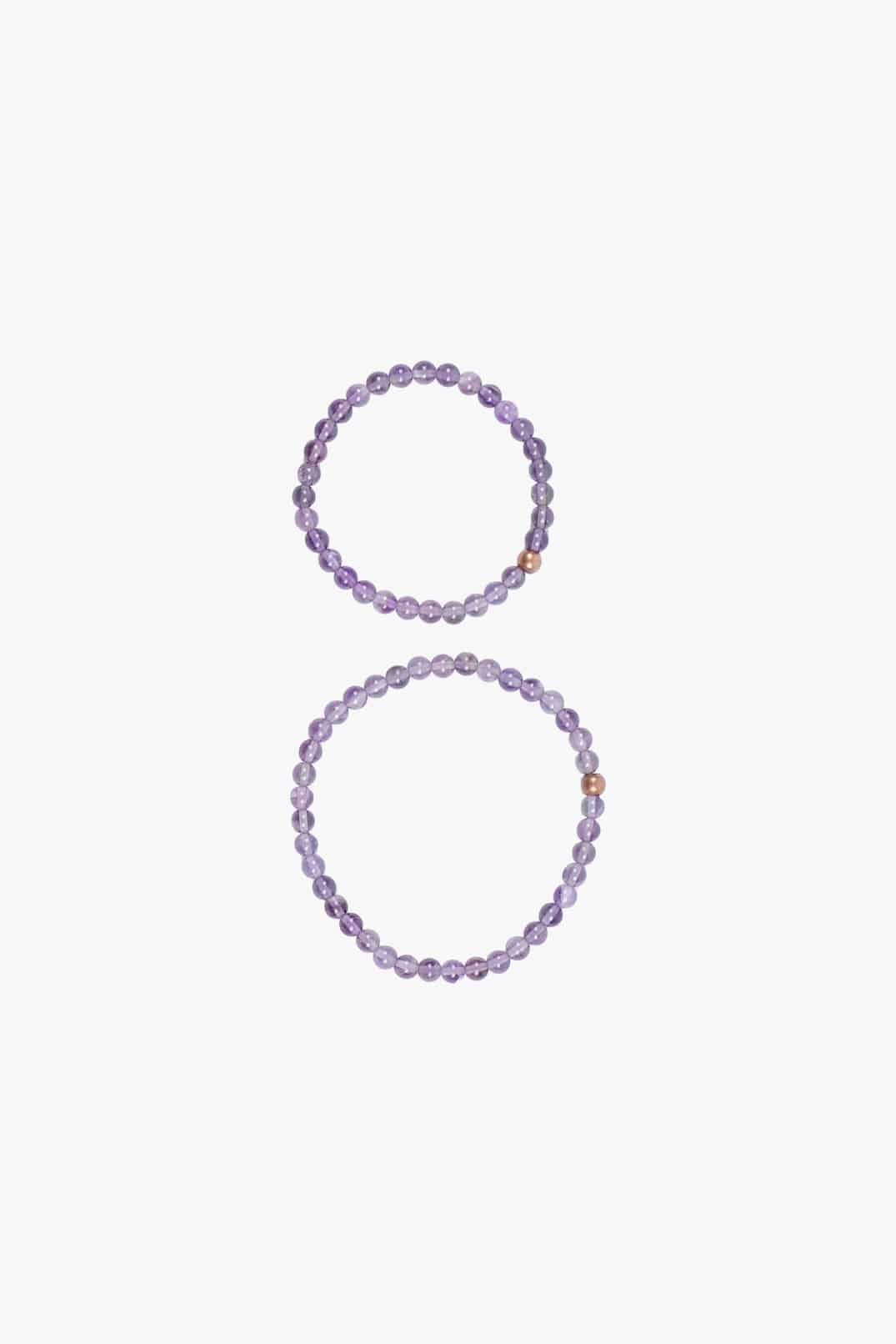 Zwei Mini und Mama Edelsteinarmbänder von Main Design aus violetten Perlen sind auf weißem Hintergrund abgebildet. Das obere Armband mit aufwendigen Amethystdetails ist etwas kleiner, während das untere größer ist. In beiden ist zwischen den violetten Perlen eine einzelne Goldperle eingearbeitet.