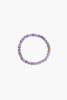 Ein Armband aus runden, polierten Amethystperlen mit einer markanten goldenen Perle. Die Mini und Mama Edelsteinarmbänder von Main Design sind in einem ordentlichen Kreis auf einem schlichten weißen Hintergrund angeordnet.