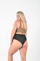 Eine Frau mit langen blonden Haaren posiert vor einem weißen Hintergrund. Sie trägt einen braunen Sport-BH und eine hoch taillierte schwarze Posh Bikinihose aus umweltfreundlichem Stoff von Main Design. Mit einem kleinen Pfeil-Tattoo auf ihrem rechten Oberarm blickt sie über ihre linke Schulter in die Kamera und sorgt so für einen nahtlos eleganten Auftritt.