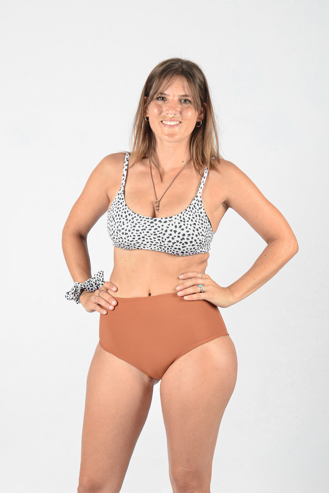 Eine Frau steht vor einem schlichten Hintergrund, lächelt und schaut in die Kamera. Sie trägt ein schwarz-weißes Bikinioberteil mit Punkten und Main Designs Posh Bikini Hose, eine hoch taillierte braune Bikinihose aus umweltfreundlichem Stoff. Sie hat ein Haargummi am Handgelenk und trägt Halsketten und Ringe für einen nahtlos stylischen Look.