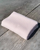 Ein ordentlich gefaltetes Baumwoll-Plaid „Soft Horizon“ von Main Design in Hellrosa mit einem dezenten Fischgrätenmuster liegt auf verwittertem Holz im natürlichen Licht.