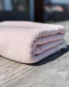 Ein gefaltetes rosa-weißes Baumwoll-Plaid „Soft Horizon“ von Main Design mit gewebtem Muster liegt auf einer verwitterten Holzoberfläche vor einem verschwommenen Außenhintergrund.