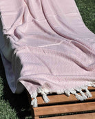 Ein hellrosa Baumwoll-Plaid „Soft Horizon“ von Main Design mit einem eingewebten Chevron-Muster und weißen Fransen ist über einen hölzernen Liegestuhl im Freien auf grünem Rasen drapiert.