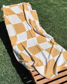 Ein hölzerner Loungesessel sitzt im Gras, drapiert mit dem schnelltrocknenden Strandtuch Sol von Main Design aus recycelter Baumwolle im gelb-weißen Karo-Muster.