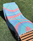 Ein Liegestuhl auf einer sonnigen Wiese ist mit dem „Main Design Strandtuch Aura“ bedeckt, einem schnell trocknenden Strandtuch mit abstrakten Mustern in Blau, Rosa und Türkis.