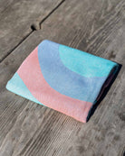 Das Strandtuch „Aura“ von Main Design aus 100 % Baumwolle besticht durch ein pastellfarbenes Muster aus blauen, rosa und türkisfarbenen geschwungenen Linien. Es trocknet schnell und eignet sich perfekt für den Strand; hier liegt es gefaltet auf einer verwitterten Holzfläche im strahlenden Sonnenlicht.