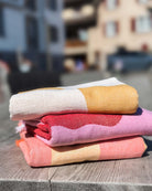 Drei gefaltete Strandtücher Sol von Main Design aus recycelter Baumwolle in Weiß, Gelb, Pink und Rot liegen auf einer Holzfläche im Freien; im Hintergrund sind unscharfe Gebäude zu sehen.