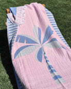 Ein Liegestuhl auf dem Rasen ist mit dem „Main Design Strandtuch Palm“ bedeckt, einem rosa-blauen Handtuch mit einem abstrakten Palmenmotiv und gestreiften Rändern.
