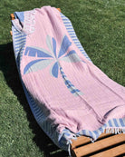 Das Main Design-Strandtuch „Palm“ mit einem rosa-blauen Palmenmotiv ist über einen Liegestuhl aus Holz auf grünem Gras drapiert, während das Sonnenlicht Schatten wirft.