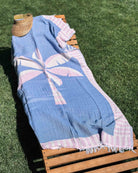 Auf einem Holzliegestuhl im Gras liegt das Strandtuch „Palm“ von Main Design in Blau-Rosa. Oben rundet ein geflochtener Sonnenhut die entspannte Sommerstimmung ab.