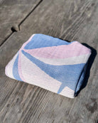 Das Strandtuch „Main Design Strandtuch Palm“, ein schnell trocknendes Strandtuch mit geometrischen Mustern in Blau, Rosa und Weiß, liegt in strahlendem Sonnenlicht auf einer verwitterten Holzfläche.