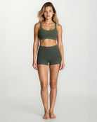 Eine Frau steht barfuß vor einem schlichten, hellen Hintergrund und trägt einen olivgrünen Sport-BH und Main Design Aura Bike Shorts mit mittlerer Leibhöhe. Sie blickt mit neutraler Miene nach vorne, die Arme liegen entspannt an ihren Seiten.