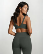 Eine Frau steht mit dem Gesicht zur Wand und trägt das Main Design Essence Active Top und hoch taillierte Leggings aus recycelten Materialien. Ihr langes gewelltes Haar fällt ihr über die Schultern, während sie vor einem schlichten hellen Hintergrund posiert.