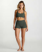 Eine Frau steht barfuß vor einem unifarbenen Hintergrund. Sie trägt das Main Design Essence Active Top und hoch taillierte Shorts aus recycelten Materialien. Sie blickt nach vorne, ein Bein leicht angewinkelt, die Arme entspannt an der Seite.