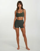 Eine Frau mit glattem braunem Haar und Pony steht barfuß vor einem hellen Hintergrund. Sie trägt das Main Design Essence Active Top und hoch taillierte Sportshorts aus recycelten Materialien, ihr Gesichtsausdruck ist neutral.