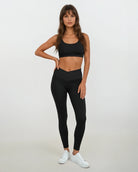 Eine Frau mit langen braunen Haaren trägt Main Design Juno Leggings mit hohem Bund aus recyceltem Nylon, einen schwarzen Sport-BH und weiße Sneakers vor weißem Hintergrund. Ihre linke Hand liegt auf der Hüfte.