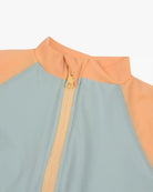 Nahaufnahme des Kai Baby Badeanzugs von Main Design mit hohem Kragen und Reißverschluss vorne. Der obere Teil des Badeanzugs ist pfirsichfarben und geht in Hellblau über. Der cremefarbene Reißverschluss aus nachhaltigen Materialien verleiht einen Hauch von Eleganz. Dieser Baby-Badeanzug bietet UV-Schutz und scheint aus glattem, dehnbarem Material zu bestehen.