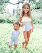 Zwei kleine Kinder stehen an einem sonnigen Tag draußen auf einer Wiese. Das ältere Kind trägt den Ava Mädchen Badeanzug von Main Design, der ein gelbes Rüschenmuster mit verstellbaren Trägern aufweist, und hält die Hand des jüngeren Kindes, das einen hellen Langarm-Badeanzug aus nachhaltigen Materialien trägt. Beide lächeln und sind barfuß.