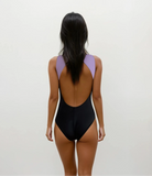 Eine Frau mit langen dunklen Haaren, von hinten gesehen, trägt den Main Design Divine Surf Badeanzug in Schwarz und Lavendel, hergestellt aus umweltfreundlichem Stoff, vor einem schlichten hellen Hintergrund.