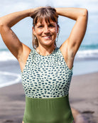 Eine lächelnde Frau steht am Strand, die Wellen des Ozeans und den Sand im Rücken. Sie trägt den Divine Surf Badeanzug von Main Design, einen nachhaltigen Badeanzug aus recycelten Materialien.