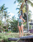 Eine Frau trägt selbstbewusst den Divine Surf Badeanzug von Main Design in Grün und Weiß, steht auf einem Baumstamm neben tropischen Palmen, während im Hintergrund klarer Himmel und Gebäude zu sehen sind.