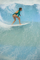 Eine Frau, die den Main Design Eclipse Surf Suit aus nachhaltigem Material trägt, surft auf einer großen blauen Welle und balanciert auf einem weißen Surfbrett, während das Wasser um sie herum plätschert.