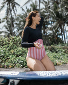 Eine Frau im Main Design Eclipse Surf Suit mit rosa und weißen Streifen und langen Ärmeln sitzt im Sand neben einem Surfbrett und lächelt mit der Hand im Haar. Hergestellt aus nachhaltigem Material für Stil und Komfort.