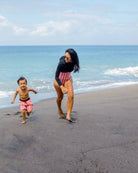 Eine Frau und ein kleines Kind, beide in passenden rot-weißen Eclipse Surf Suits von Main Design aus nachhaltigem Material, rennen und lächeln an einem Sandstrand in der Nähe des Ozeans unter einem teilweise bewölkten Himmel.