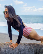 Mit dem Main Design Eclipse Surf Suit aus nachhaltigem Material mit UPF-Schutz macht eine Frau eine Planke auf Treibholz am Strand, umrahmt von Wellen und einem bewölkten Himmel.