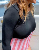 Eine Frau im schwarz-rosa gestreiften und aus nachhaltigen Materialien gefertigten Eclipse Surf Suit von Main Design steht auf einem Surfbrett am Strand mit dem Meer im Hintergrund.