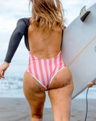 Eine Frau im rosa-weiß gestreiften und aus nachhaltigem Material gefertigten Eclipse Surf Suit von Main Design geht mit sandigen Beinen und einem Surfbrett unter dem Arm auf den Ozean zu.