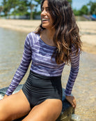 Eine Frau mit langen, welligen Haaren sitzt lächelnd am Wasserrand eines Strandes. Sie trägt einen lilafarbenen Batik-Surfanzug „Ivy“ von Main Design aus recycelten Materialien und eine hoch taillierte schwarze Hose. Im Hintergrund sind Bäume und ein heller Himmel zu sehen.