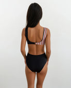 Eine Frau blickt von der Kamera weg und trägt den Luna Badeanzug von Main Design – ein hoch tailliertes schwarzes Bikinihöschen und ein gestreiftes One-Shoulder-Top aus recycelten Materialien – vor einem schlichten hellen Hintergrund.