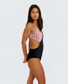 Eine Frau steht im Profil und trägt den Luna Badeanzug von Main Design mit rosa und weißen Streifen und einem einfarbig schwarzen Unterteil mit seitlichem Ausschnitt – gefertigt aus recycelten Materialien für Eleganz und Funktionalität vor einem schlichten Hintergrund.