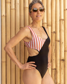 Eine lächelnde Frau mit kurzen grauen Haaren und Sonnenbrille trägt den Luna Badeanzug von Main Design – einen einteiligen Badeanzug mit rosa Streifen, schwarzen Abschnitten und dezenten Ausschnitten – und posiert selbstbewusst vor einer Bambuswand.