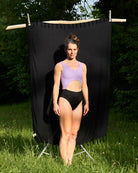 Eine Person, die den aus recycelten Materialien hergestellten Nova Badeanzug von Main Design trägt, steht auf Gras vor einem schwarzen Stoffhintergrund inmitten von grünen Bäumen und Sonnenlicht.